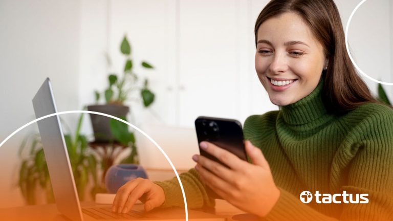 Imagem mostra jovem mulher sorridente mexendo no celular e no notebook ao mesmo tempo, de blusa verde, para ilustrar como ganhar dinheiro na Kiwify