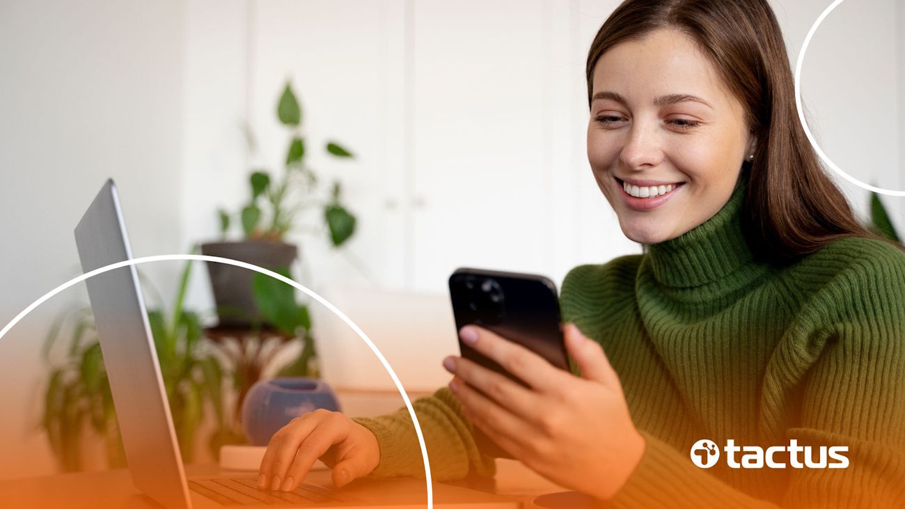 Imagem mostra jovem mulher sorridente mexendo no celular e no notebook ao mesmo tempo, de blusa verde, para ilustrar como ganhar dinheiro na Kiwify