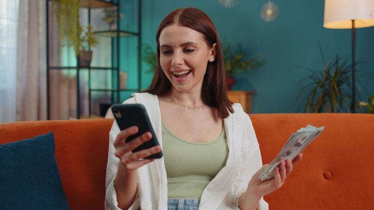 Imagem mostra jovem sorrindo e segurando dólares na mão junto com celular, para ilustrar como ganhar dinheiro no Instagram