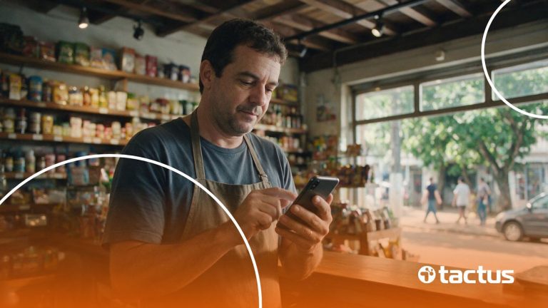 Pequeno comerciante de meia idade em sua loja olhando para o celular, para ilustrar as mudanças da reforma tributária para o MEI