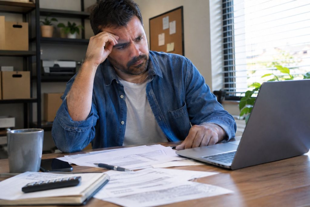 pequeno empresário brasileiro sentado à mesa de trabalho com papéis espalhados, olhando para o notebook com expressão preocupada querendo regularizar cnpj
